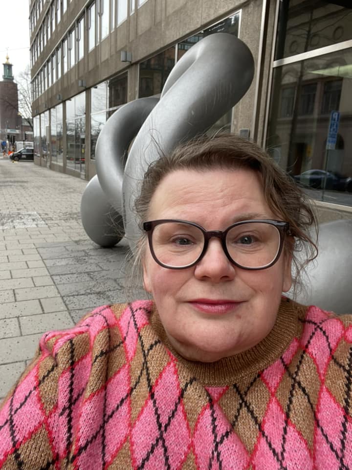 Maria på trottoaren utanför regeringskansliets lokaler på Jakobsgatan. Bakom henne syns skulpturen Binär, av Eva Hild. Maria är klädd i en tröja med rosa/röda romber mot en ljusbrun bakgrund.